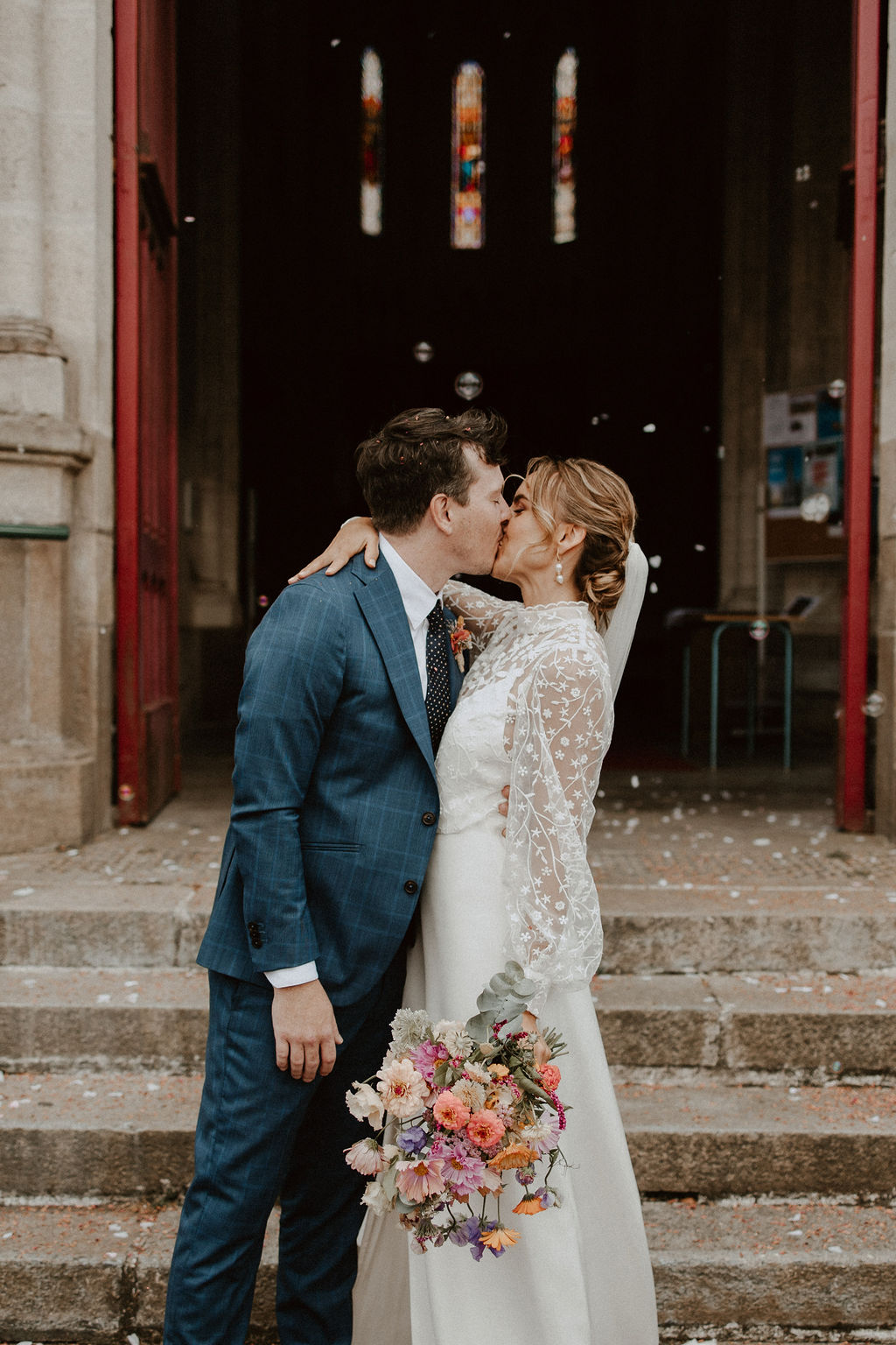 Alice et Clément s’embrassent tendrement devant leurs invités, sous une pluie de pétales de fleurs, dans un cadre champêtre et romantique.